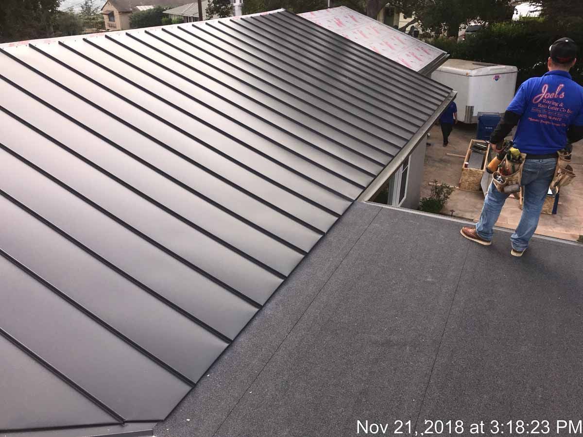Roofers working on a dark metal roof beside a flat roof, with tools and materials visible.