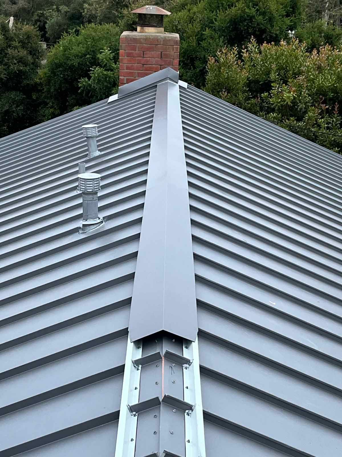 Metal roof ridge with chimney, vents, and a valley against green trees