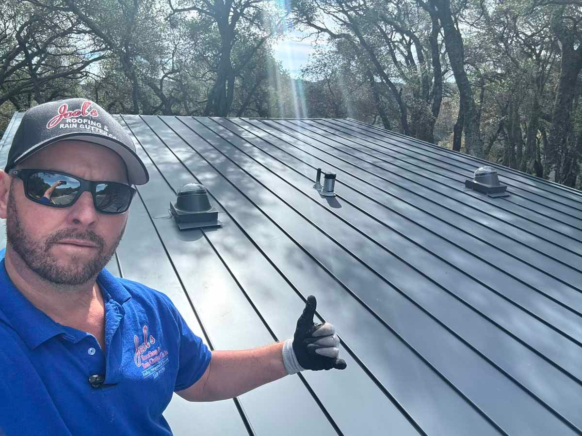 Man in sunglasses giving a thumbs-up on a dark metal roof with trees in the background