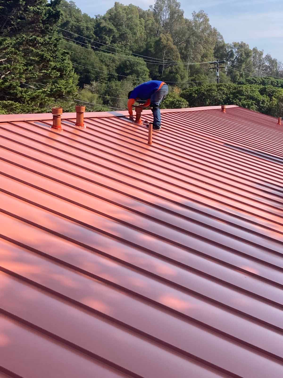 Worker on a red metal roof using a harness, with trees in the background.