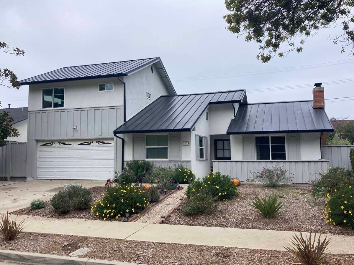 Two-story house with dark metal roof, white exterior, and desert landscaping under an overcast sky