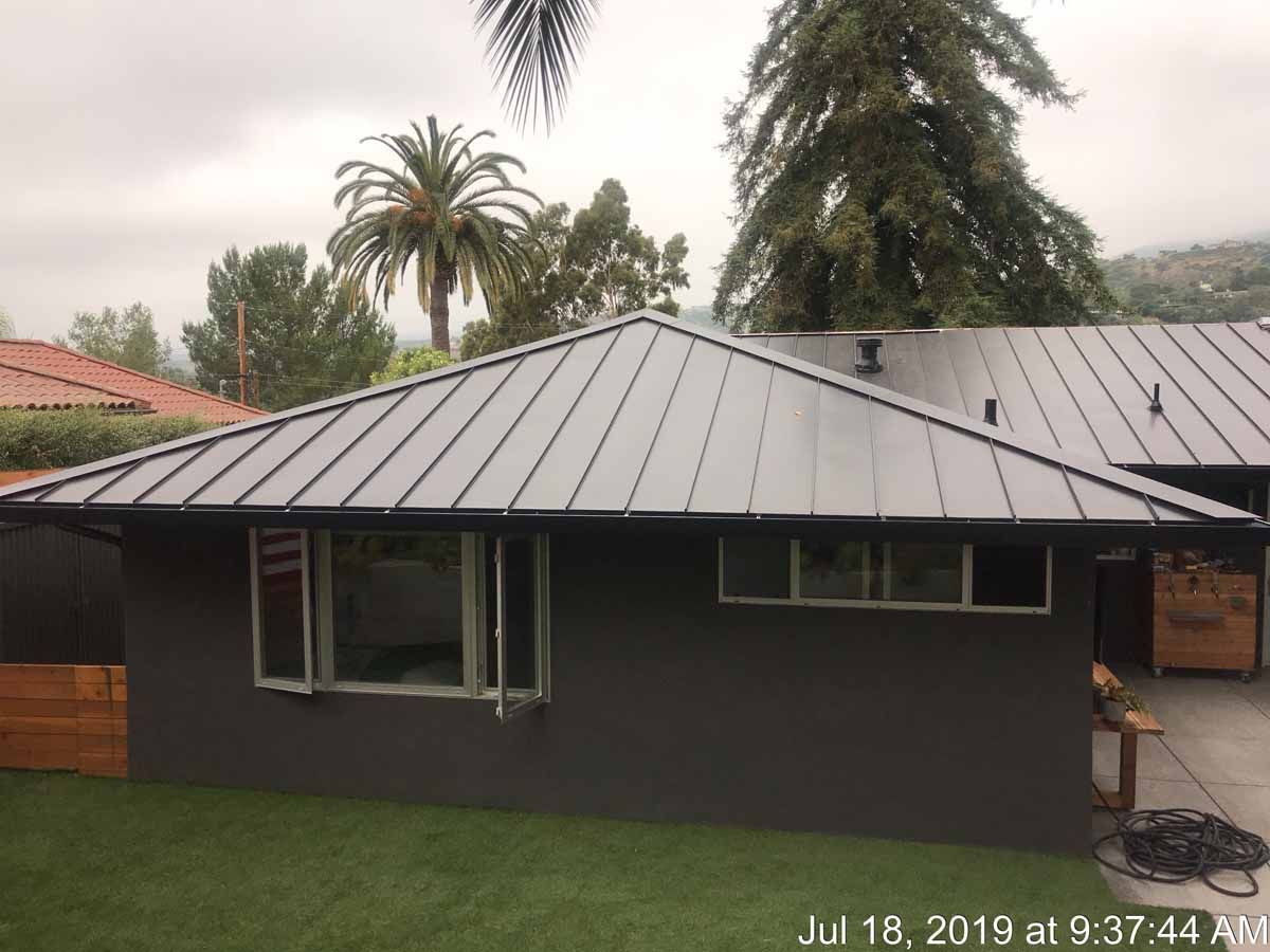 Dark gray house with a metal roof in a rainy suburban yard, palm trees in the background