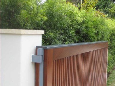 Wooden slat driveway gate beside a white wall, with green bushes behind it