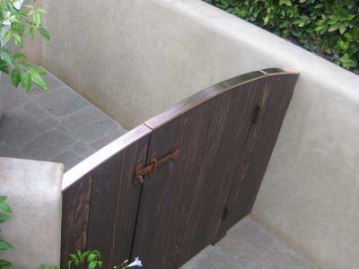 A dark wooden gate with a curved top set into a light stone wall beside greenery.