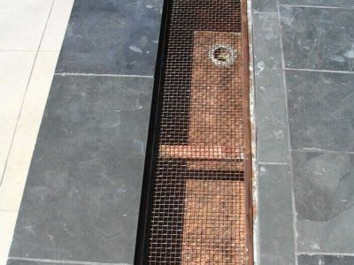 Narrow drainage channel with metal grating between gray and white floor tiles, with a circular drain cover