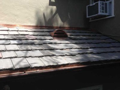 Snow-covered shingled roof with a vent and an air conditioner window unit on the side wall.