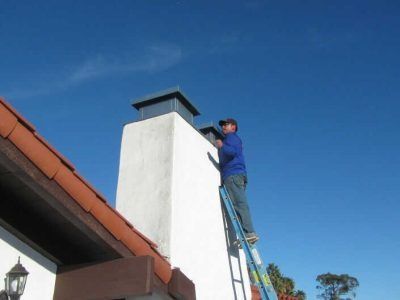 Person on a ladder painting a white chimney on a house roof under a clear blue sky