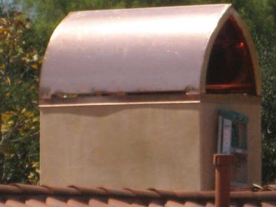 Small rooftop structure with a curved metallic roof and tan walls against trees and sky.
