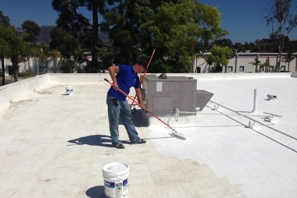Worker painting a flat white rooftop near HVAC equipment and a bucket of paint