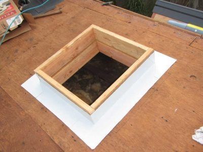 Square wooden roof hatch on a plywood surface with white flashing around the opening