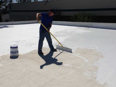 Person using a squeegee to apply white coating on a flat rooftop beside a paint bucket