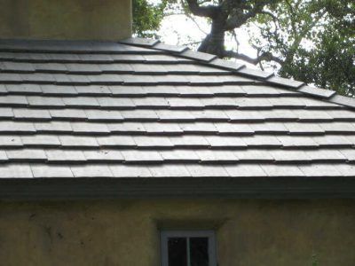 Close-up of a gray shingled roof with a small window below and tree branches above