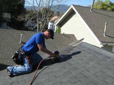 Worker on a roof using a power tool to repair gray shingles near a skylight.