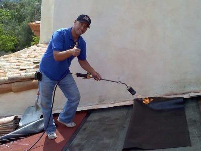 Worker using a torch to heat roofing material on a flat rooftop, wearing blue shirt and cap