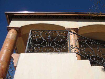 Low-angle view of a house balcony with ornate metal railing and cream-colored columns against a blue sky