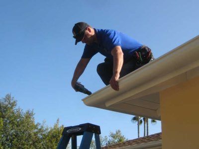 Worker in blue shirt kneeling on a roof edge, installing a gutter under a clear blue sky.