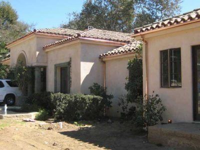 Single-story beige stucco house with tile roof, shrubs, and a partial white vehicle in the driveway