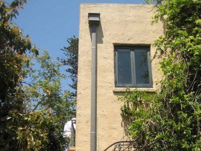 Cream-colored building wall with a window and gutter, framed by green trees against a blue sky