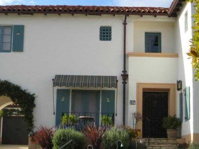 White stucco building with green shutters, a striped awning, and potted plants at the entrance