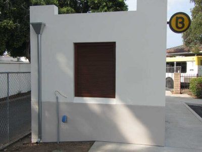 White utility building with a brown shuttered window and a yellow “B” sign outdoors