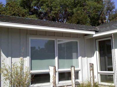 Single-story house exterior with gray siding, two windows, and a small front porch under trees.