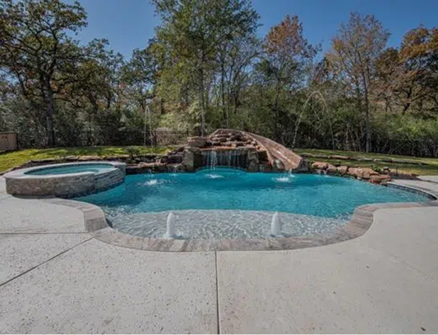 Swimming pool with waterfall, hot tub, surrounded by concrete patio and trees.