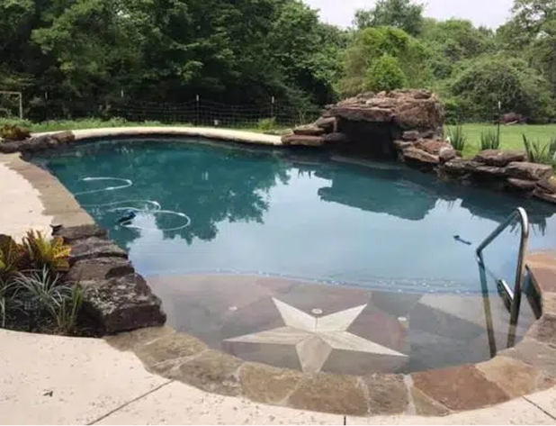 Swimming pool with Texas star design, rock waterfall, and surrounding trees.