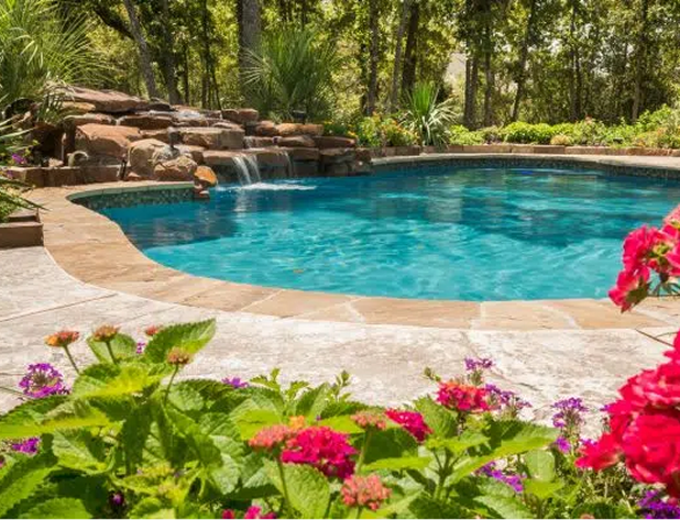 Swimming pool with waterfall feature, surrounded by landscaping, with trees in the background.
