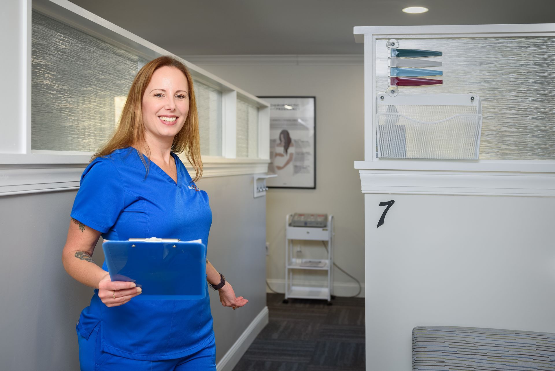 A woman in a blue scrub is standing in a hallway holding a clipboard.Touch