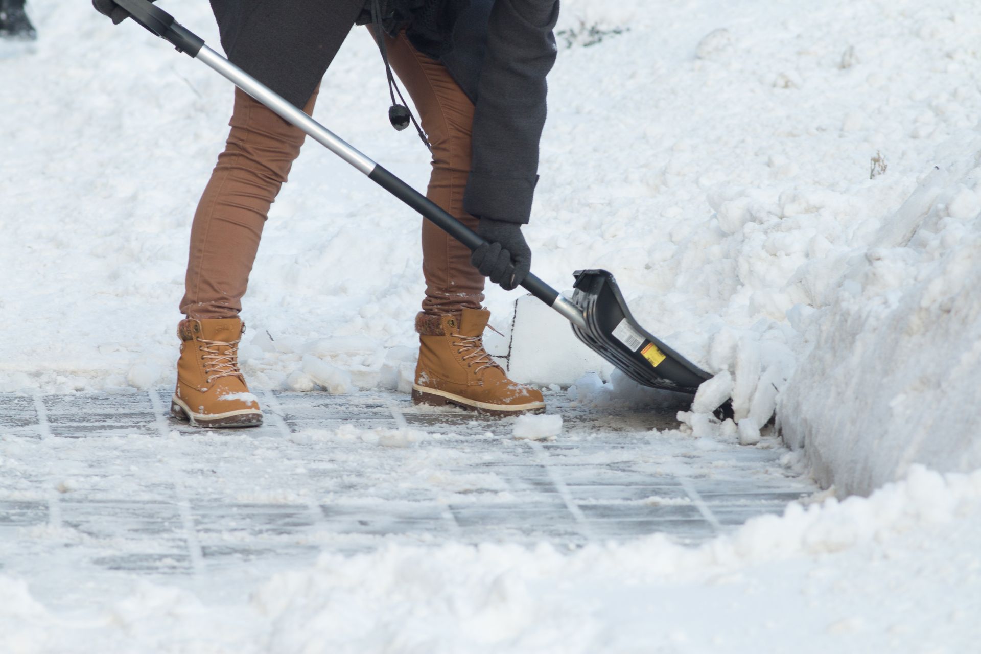 Person shoveling snow from a brick walkway; brown pants, boots, and black jacket; gray shovel.