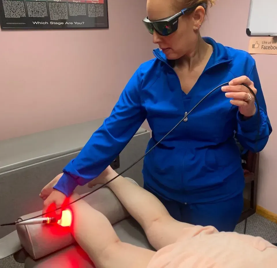 A woman in a blue jacket is using a red light on a person 's leg