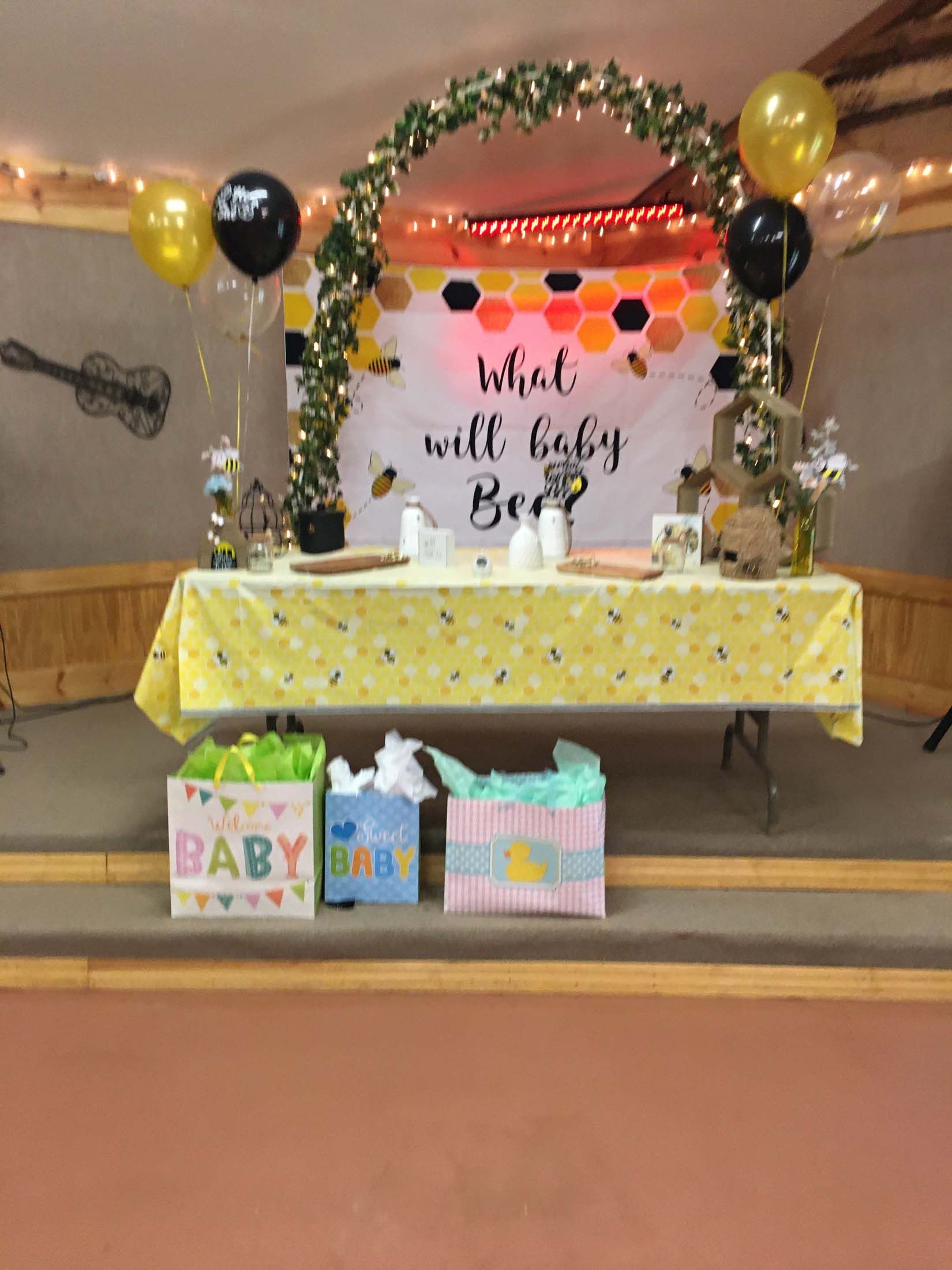 Baby shower table with yellow and black balloons, a sign, and gift bags below the table