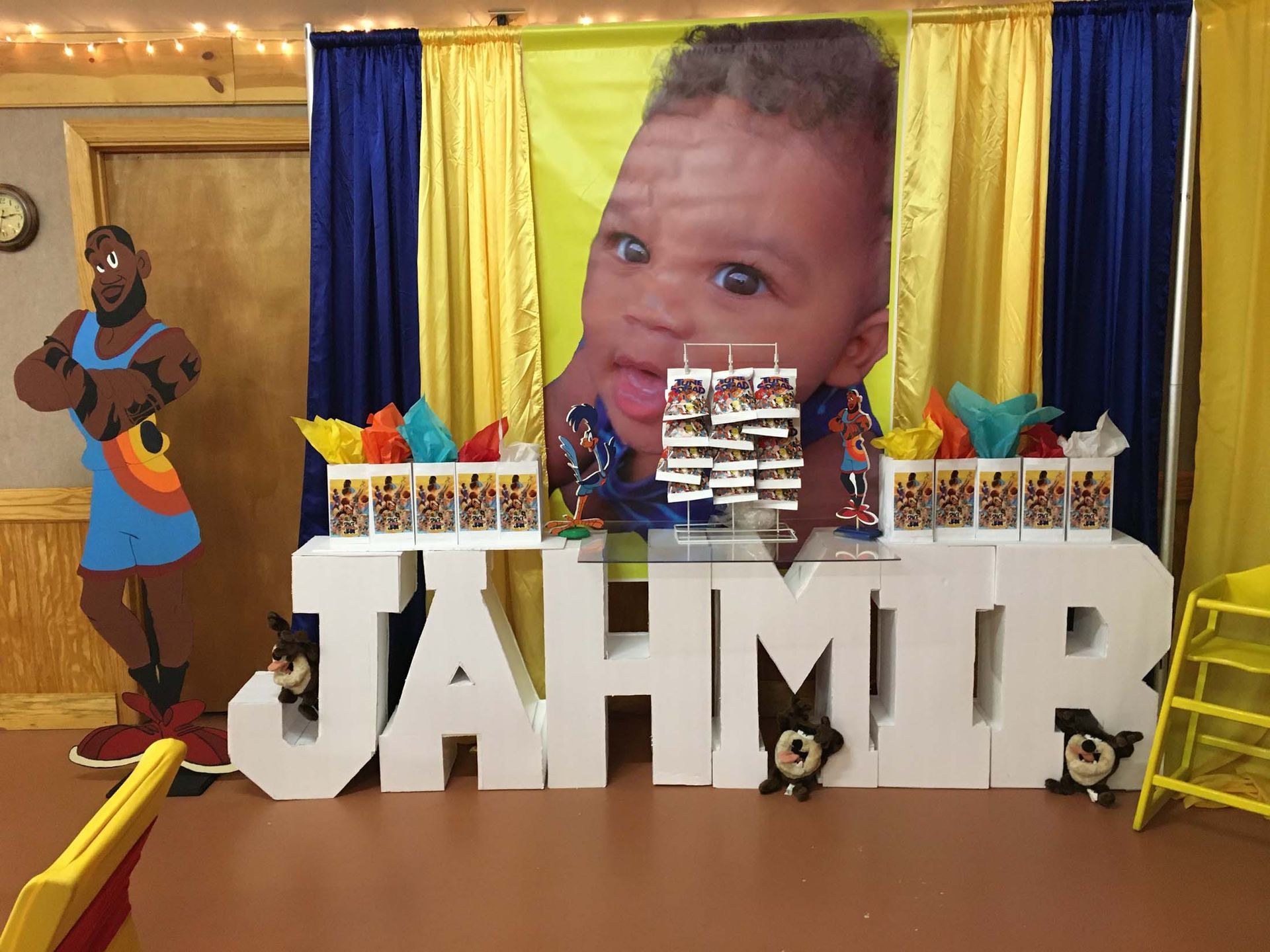 Decorated party backdrop with large “JAHMIR” letters, colorful drapes, balloons, and a baby photo banner