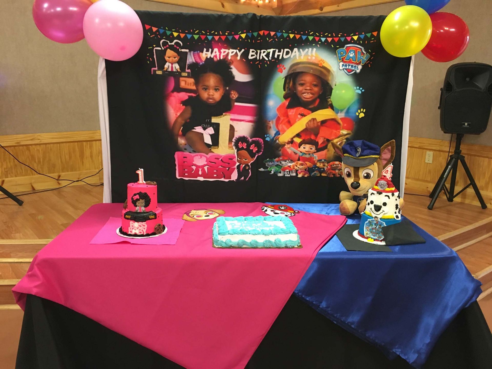 Birthday party table with colorful cake, balloons, and a “Happy Birthday” backdrop of two children.