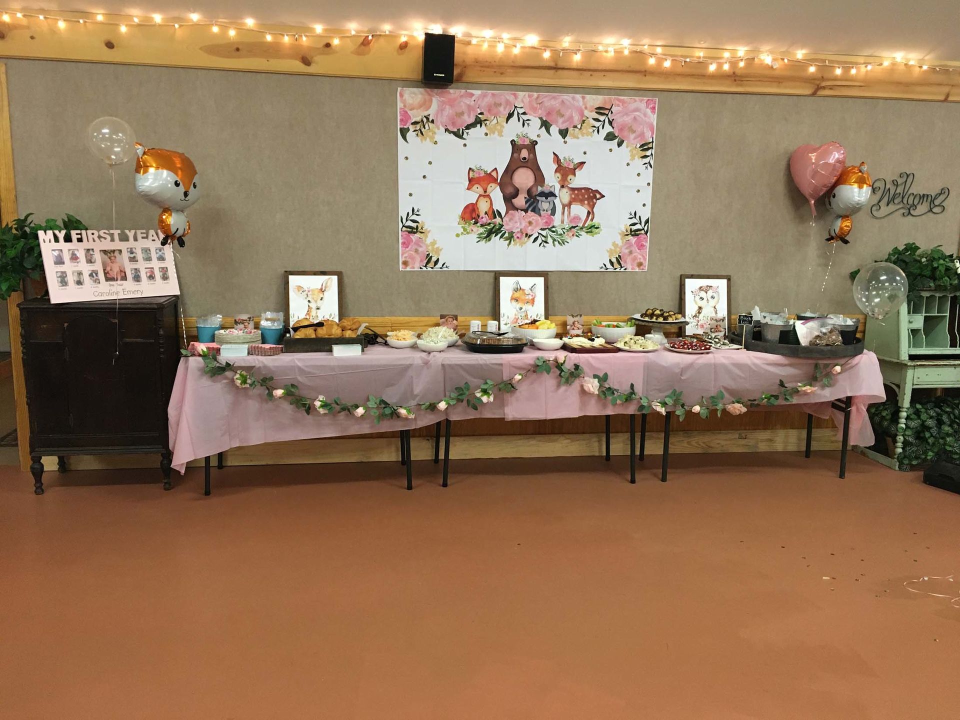 Decorated dessert buffet table with pink cloth, fairy lights, balloons, and pastel cartoon banner for a celebration