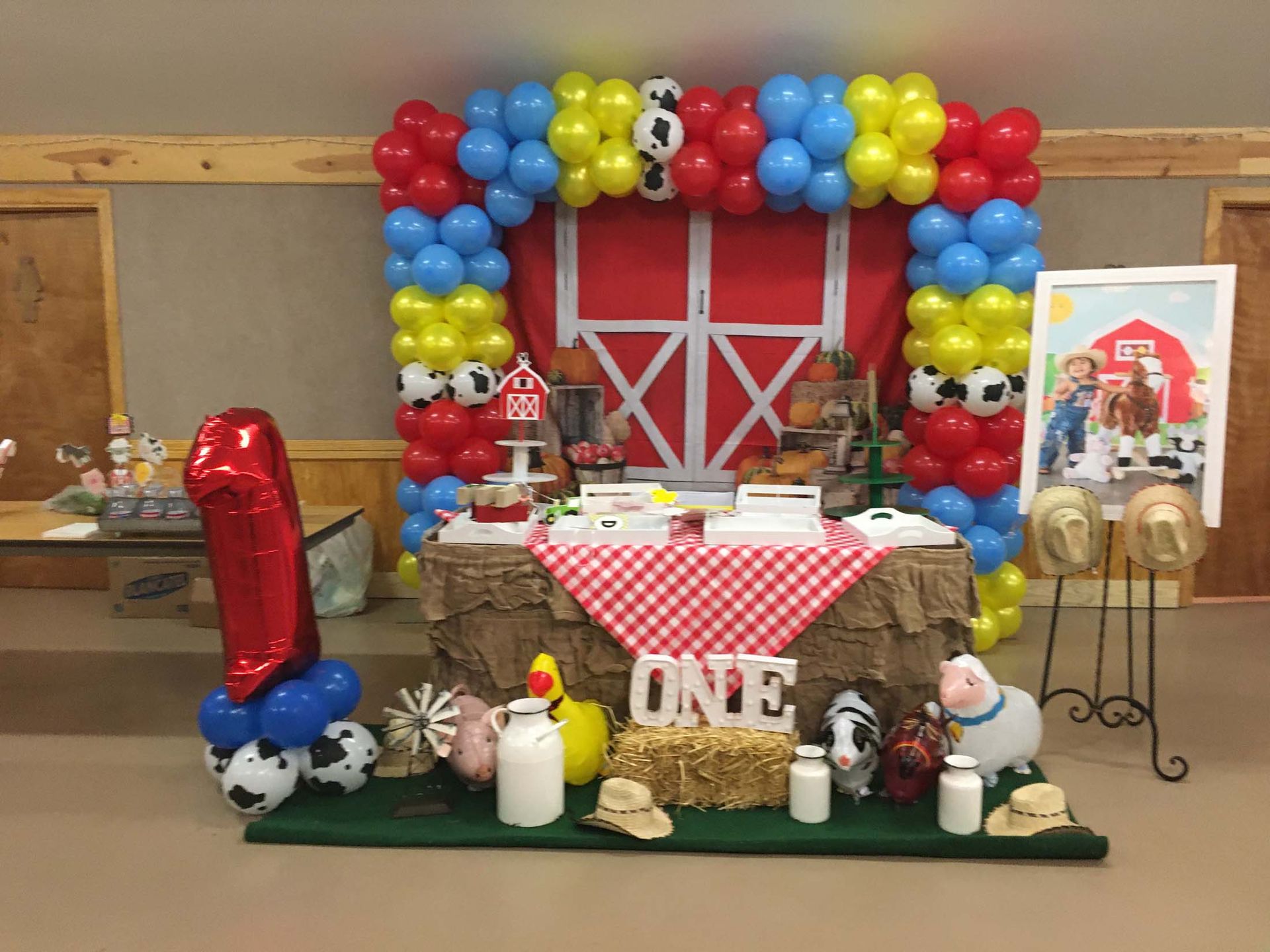 Balloon-decorated barn-themed party table with gifts and stuffed animals, red-and-blue balloons, and a “ONE” sign