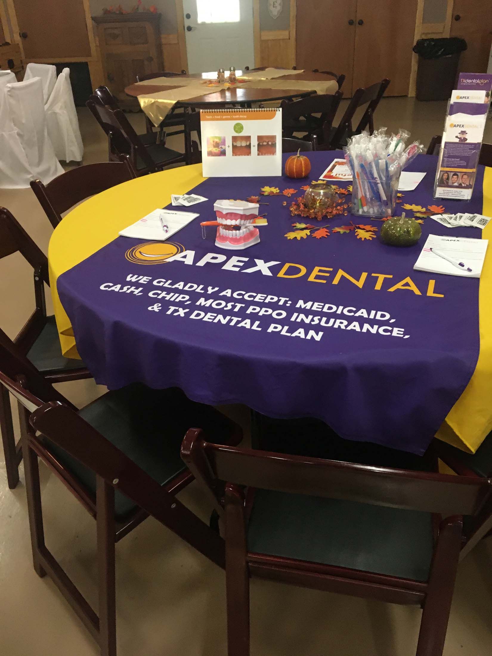 Dental outreach table with purple “APEX DENTAL” cloth, brochures, and supplies in a room