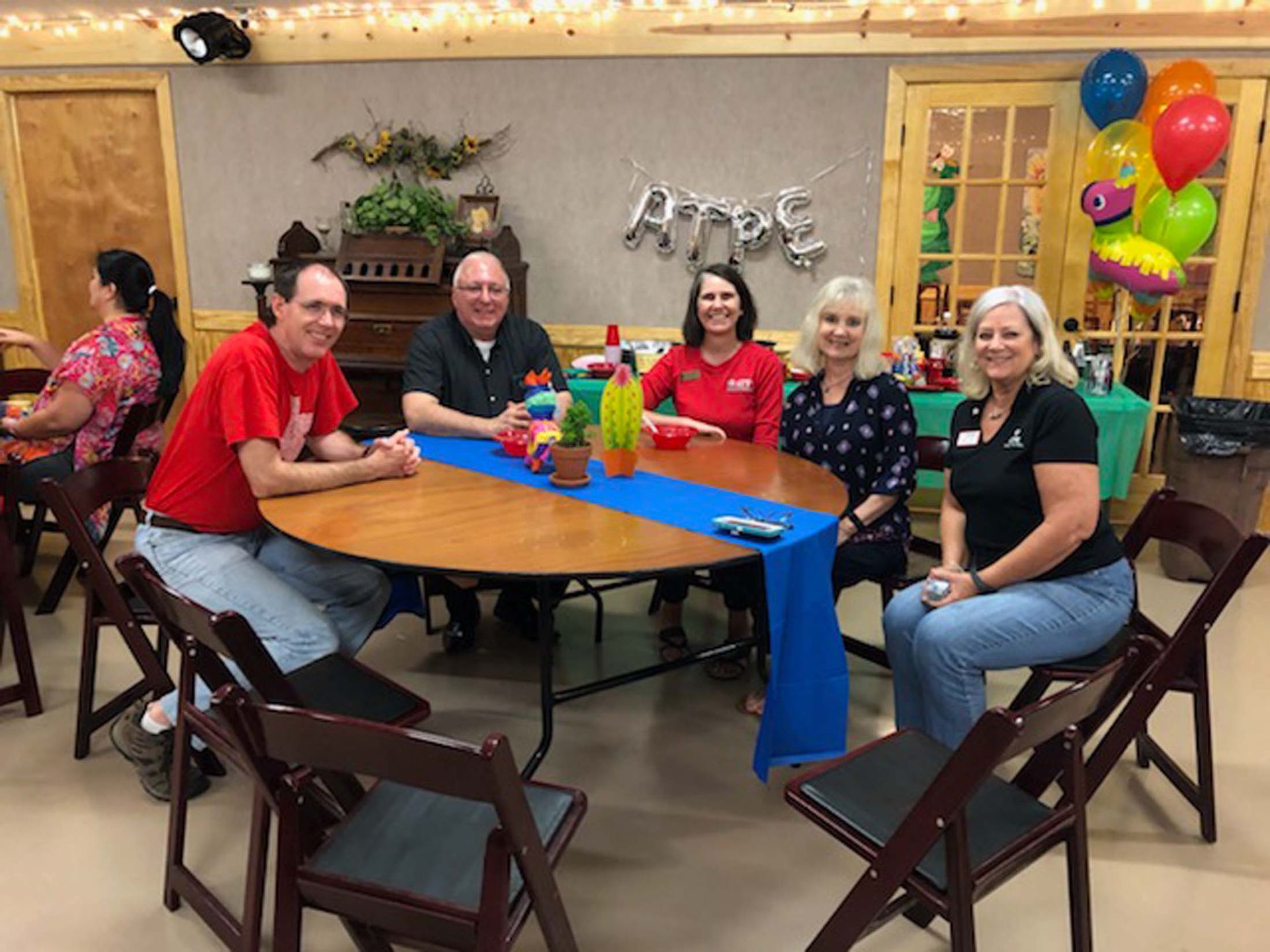 Six people seated around a table at a festive indoor gathering with balloons and decorations.