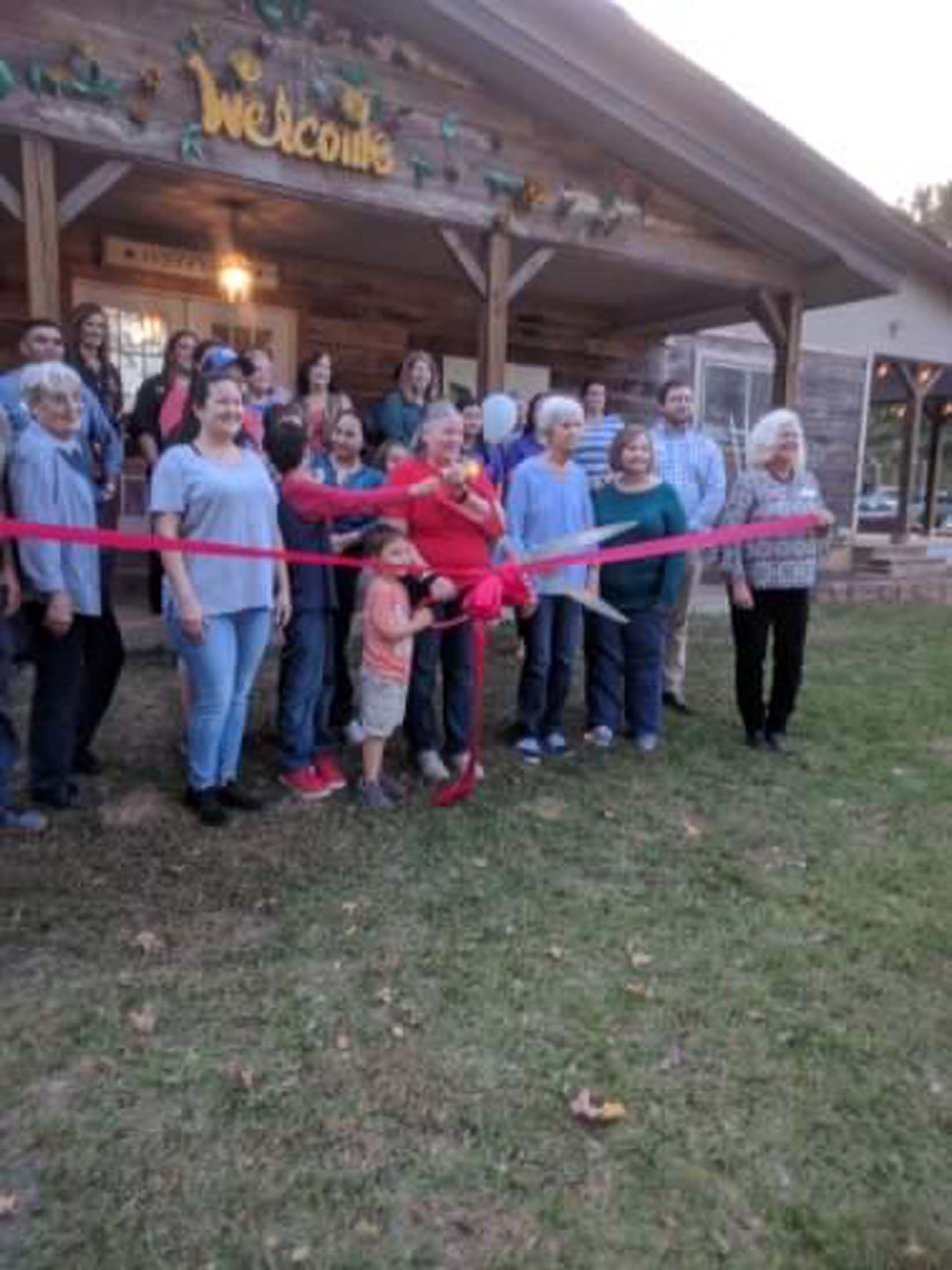 Group posing outside a wooden building at a ribbon-cutting ceremony, with a red ribbon across the front.