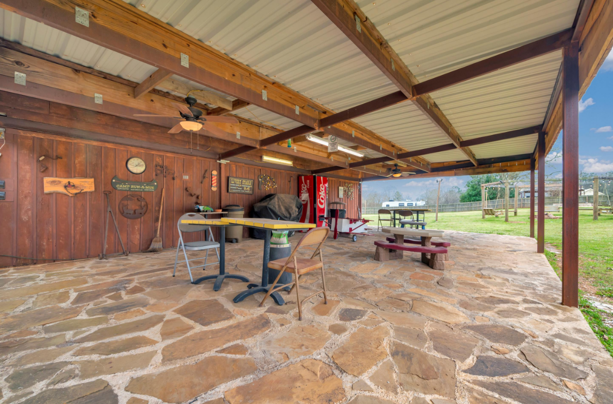 Covered stone patio with picnic table, benches, and wooden wall under a metal roof