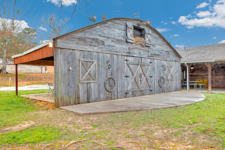 Weathered gray barn with double doors on a concrete pad, set in a grassy rural yard under a blue sky