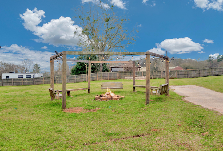 Wooden pergola over a grassy park area with a fire pit and a paved path under a blue sky.