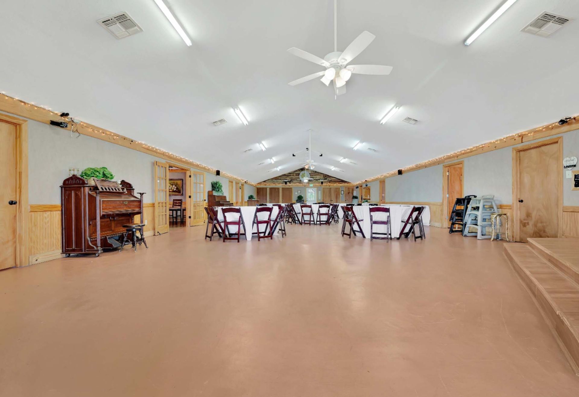 Spacious hall with tan floor, white ceiling fans, chairs, and tables arranged at the far end