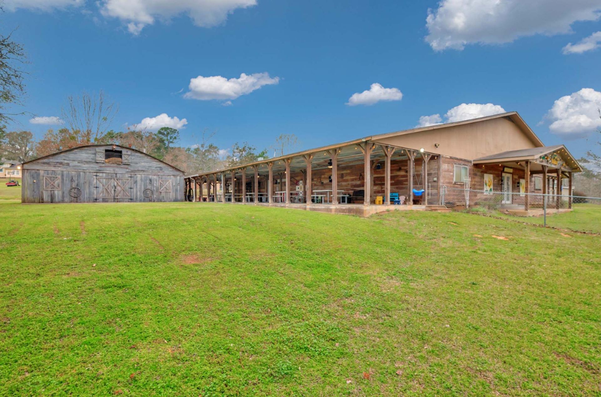 Rural farmyard with a large wooden barn, stone shed, and grassy field under a blue sky
