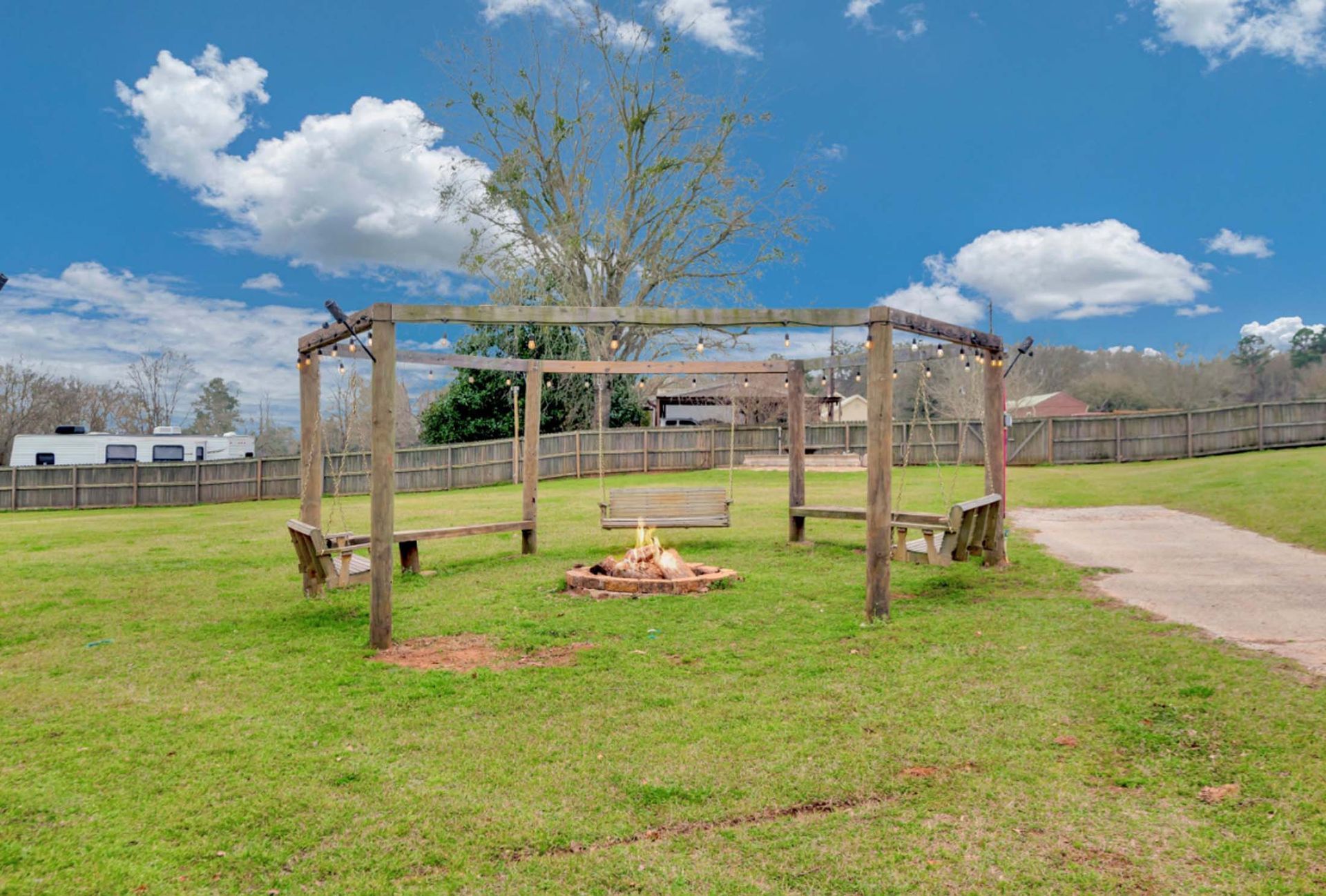 Wooden pergola over a grassy park area with a fire pit and a paved path under a blue sky.