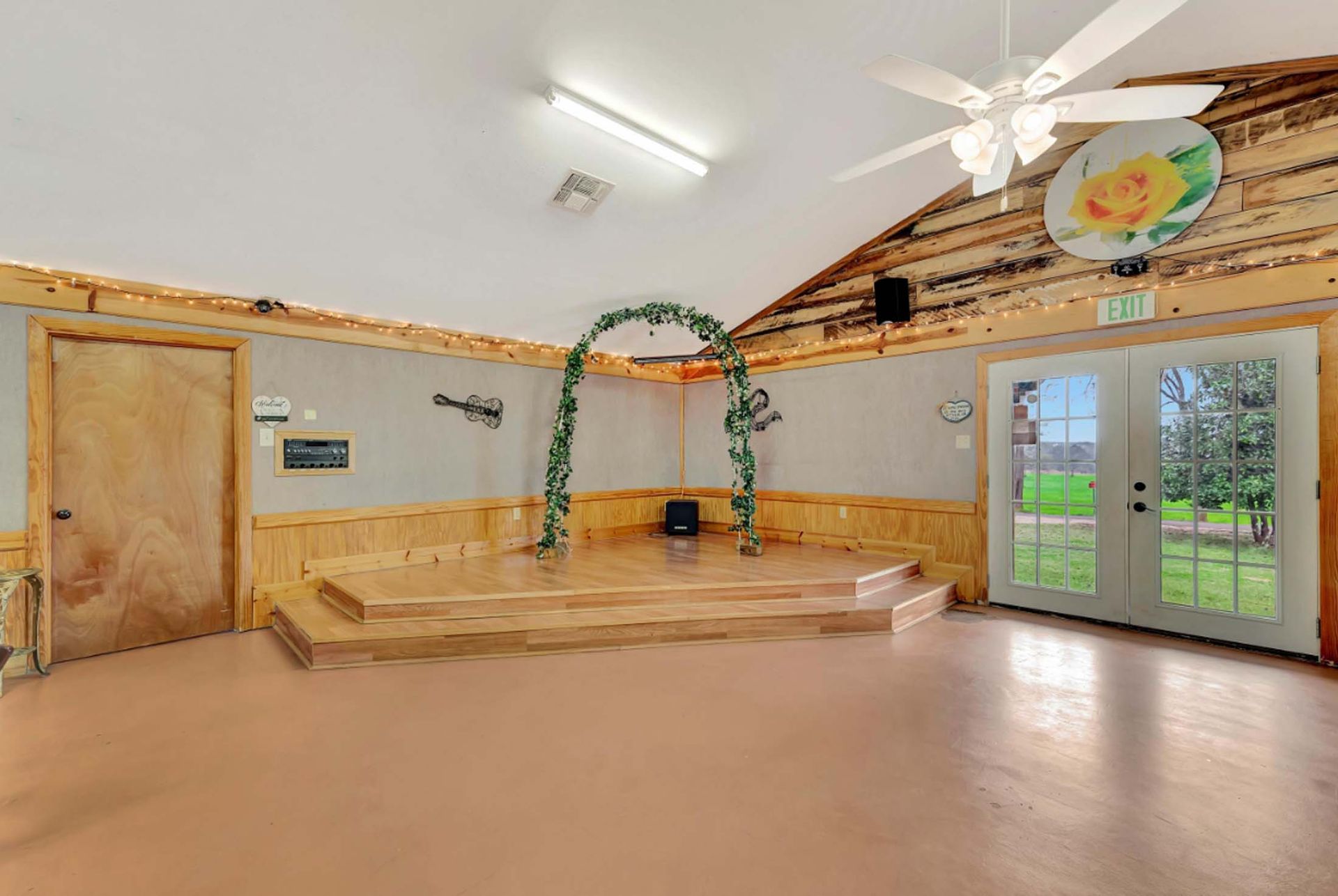 Bright indoor hall with wood trim, small stage, archway, and glass doors opening to greenery