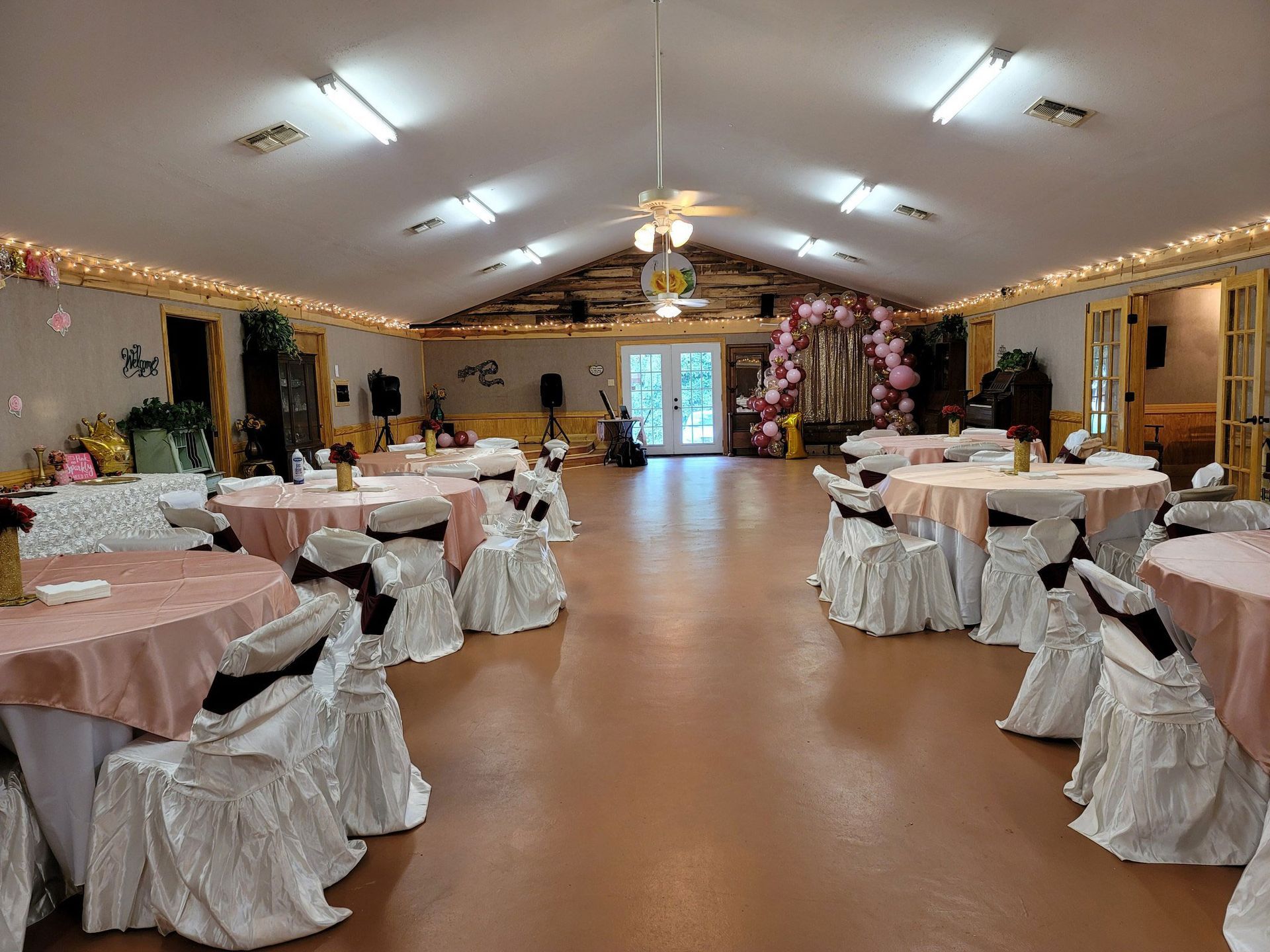 Decorated banquet hall with round tables, pink tablecloths, and white chair covers under bright ceiling lights