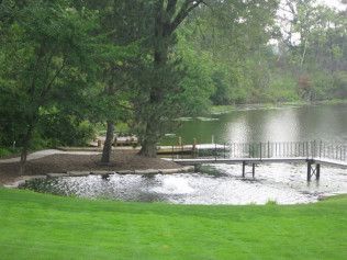 Lush green lawn slopes to a pond with a small pier and a fountain. Trees frame the water's edge.