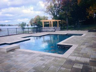 Outdoor pool with pavers, overlooking water and gazebo with trees under a cloudy sky.