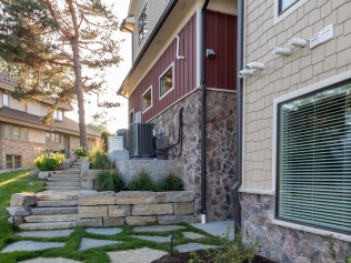 Stone steps and path lead to a building with stone and tan siding. Landscaping surrounds.